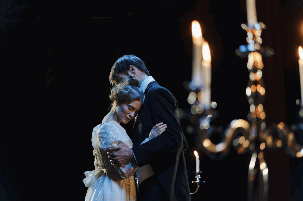 two actors are on stage in an embrace. the background is dark and there is a candelabra in the foreground.